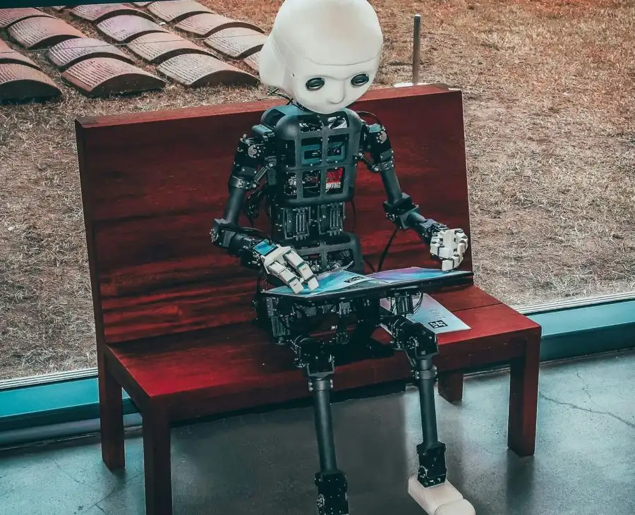 Humanoider Roboter sitzt auf einer Holzbank und liest eine Broschüre — Symbolbild zur KI-Kennzeichnungspflicht im Beitrag von quirkativ